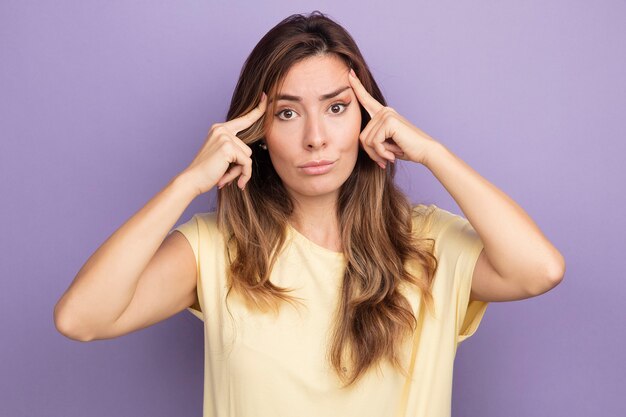 Young beautiful woman in beige t-shirt looking at camera displeased touching her temples