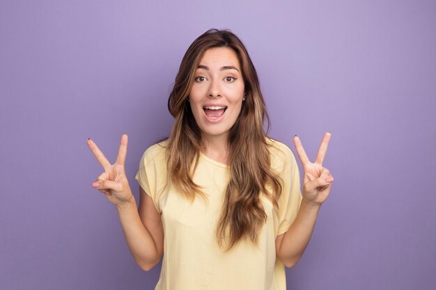 베이지 색 t- 셔츠에 젊은 아름 다운 여자 카메라 행복 하 고 보라색 위에 서있는 v 기호를 보여주는 흥분