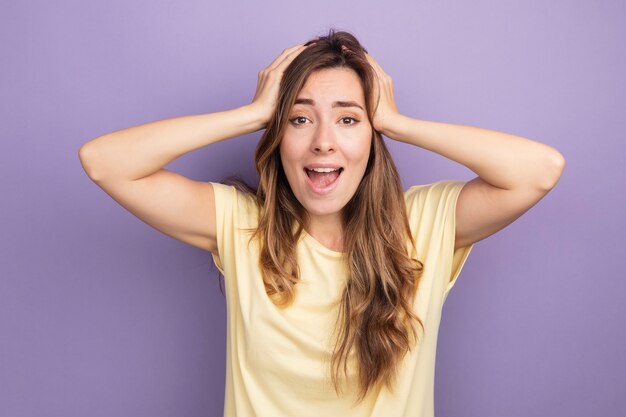 Young beautiful woman in beige t-shirt looking at camera happy and excited with hands on her head standing over purple background
