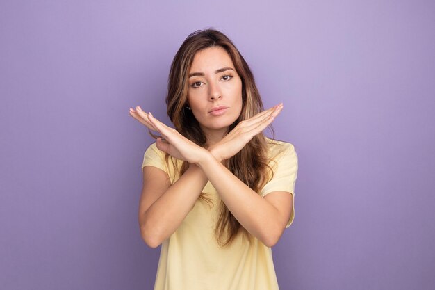 Young beautiful woman in beige t-shirt looking at camera with serious face making stop gesture crossing hands standing over purple background