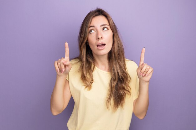 Young beautiful woman in beige t-shirt looking up surprised pointing with index figners up standing over purple