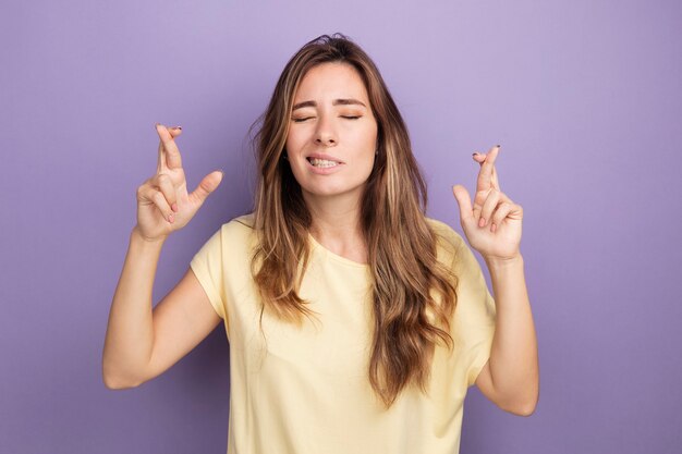 Young beautiful woman in beige t-shirt making desirable wish with closed eyes crossing fingers standing over purple