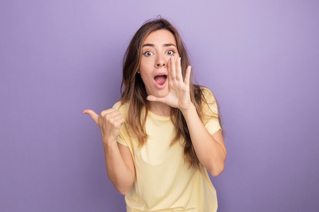 Молодая красивая женщина в бежевой футболке кричит рукой возле рта, указывая назад с большим пальцем, стоящим на фиолетовом фоне