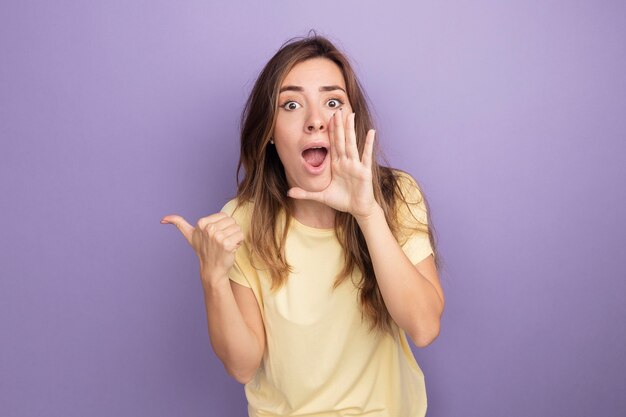 Young beautiful woman in beige t-shirt shouting with hand near mouth pointing back with thumb standing over purple background