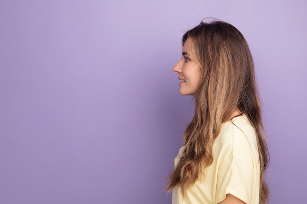 Foto gratuita giovane bella donna in maglietta beige in piedi di lato con un sorriso sul viso su sfondo viola