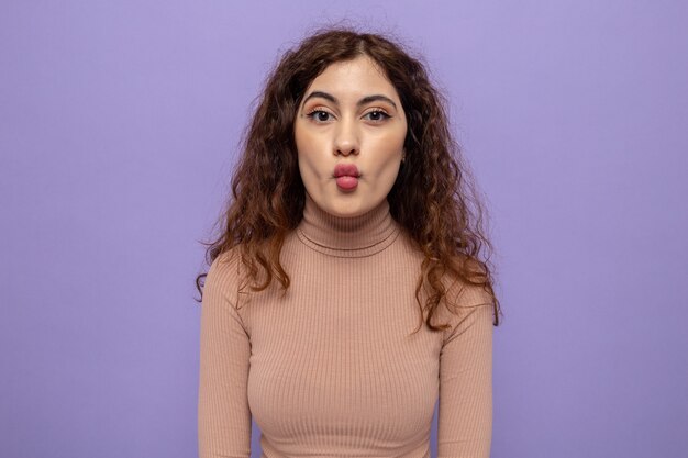 Young beautiful woman in beige turtleneck  keeping lips as going to kiss standing over purple wall