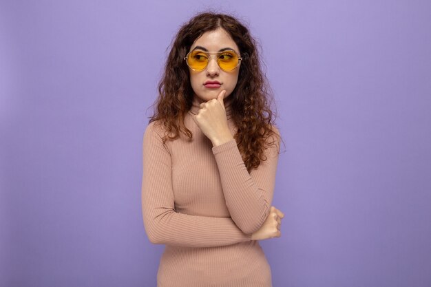 Young beautiful woman in beige turtleneck wearing yellow glasses looking aside with hand on her chin with pensive expression
