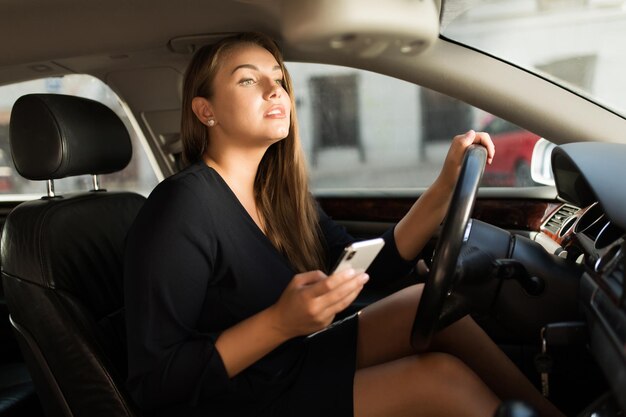 思慮深くまっすぐ見ながら携帯電話を手に持って車を運転する車輪の後ろに座っている黒いドレスを着た若い美しい女性