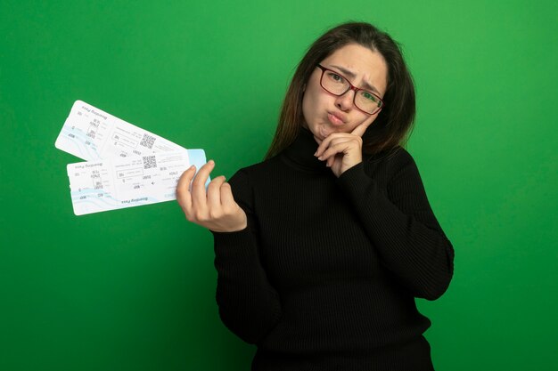 Foto gratuita giovane bella donna in un dolcevita nero e occhiali in possesso di biglietti aerei guardando la parte anteriore confusa e scontenta in piedi sopra la parete verde