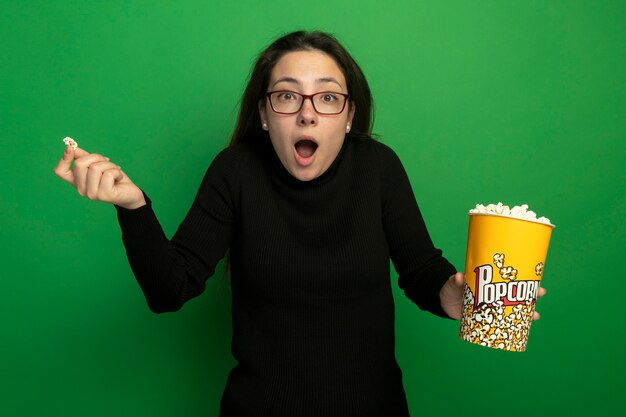 Young beautiful woman in a black turtleneck and glasses holding bucket with popcorn looking confused with raised arm standing over green wall