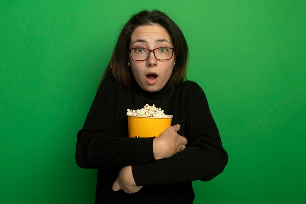 Young beautiful woman in a black turtleneck and glasses holding bucket with popcorn looking at front with fear expression standing over green wall