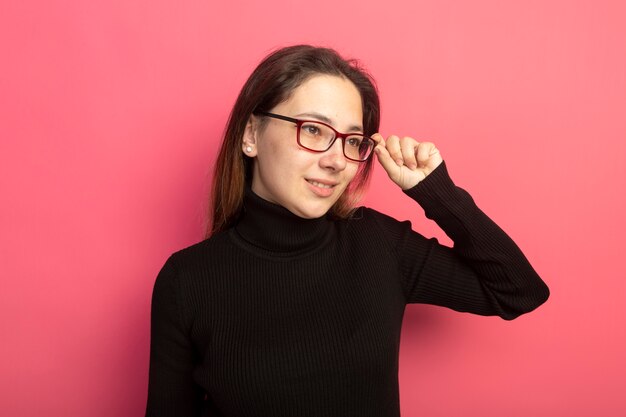 Young beautiful woman in a black turtleneck and glasses looking aside with smile on face touching her glasses standing over pink wall