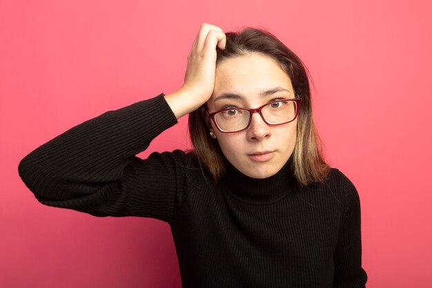 Young beautiful woman in a black turtleneck and glasses looking at front puzzled standing over pink wall