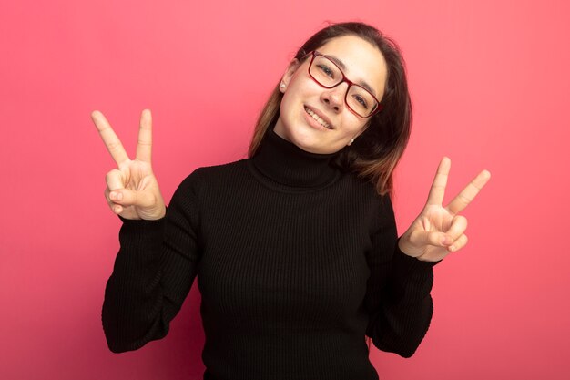 Young beautiful woman in a black turtleneck and glasses looking at front smiling cheerfully showing v-sign standing over pink wall