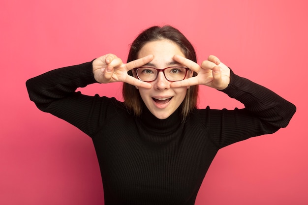 ピンクの壁の上に立っている彼女の目の近くにVサインを示す幸せそうな顔で笑顔で正面を見て黒いタートルネックとメガネの若い美しい女性