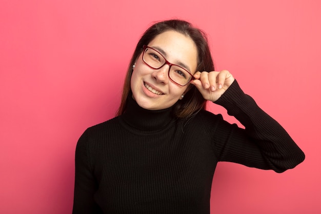 Young beautiful woman in a black turtleneck and glasses looking at front smiling with happy face touching her glasses standing over pink wall