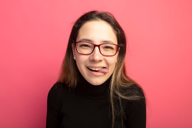 Young beautiful woman in a black turtleneck and glasses looking at front sticking out tongue happy and positive standing over pink wall