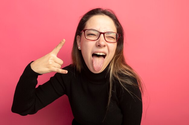 Young beautiful woman in a black turtleneck and glasses looking at front sticking out tongue making rock symbol standing over pink wall