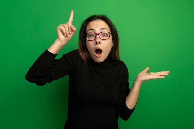 Young beautiful woman in a black turtleneck and glasses looking at front surprised and amazed showing index finger presenting something with arm standing over green wall