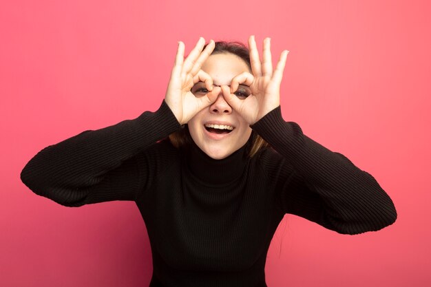 Foto gratuita giovane bella donna in un dolcevita nero e occhiali guardando davanti attraverso le dita che fanno il gesto binoculare in piedi sopra la parete rosa