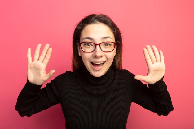 Young beautiful woman in a black turtleneck and glasses looking at front with raised hands standing over pink wall