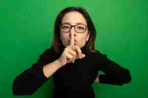 Free photo young beautiful woman in a black turtleneck and glasses, making silence gesture with finger on lips standing over green wall