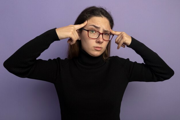 Young beautiful woman in a black turtleneck and glasses pointing with fingers at her temples trying to concentrate on a task standing over purple wall