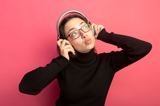 Giovane bella donna in un dolcevita nero e occhiali con le cuffie che osserva in su in piedi felice e positivo sopra il muro rosa