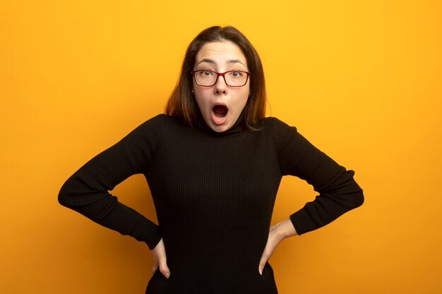 Young beautiful woman in a black turtleneck looking at front being amazed and surprised standing over orange wall