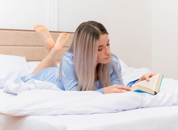 침실 인테리어에 독서 책을 쉬고 침대에 누워 파란색 잠옷에 젊은 아름 다운 여자