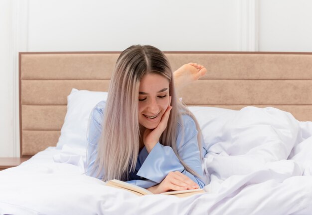 Young beautiful woman in blue pajamas laying on bed resting reading book smiling in bedroom interior 
