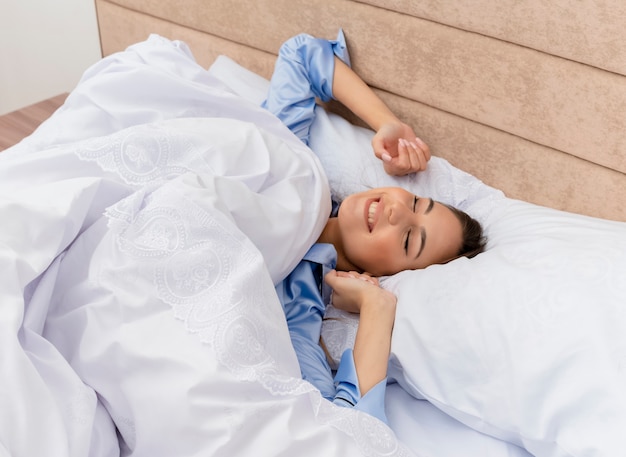 Free photo young beautiful woman in blue pajamas laying on bed resting on soft pillows waking up stretching her hands in bedroom interior on light background