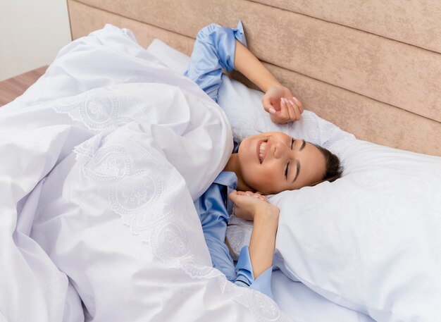 Young beautiful woman in blue pajamas laying on bed resting on soft pillows waking up stretching her hands in bedroom interior on light background