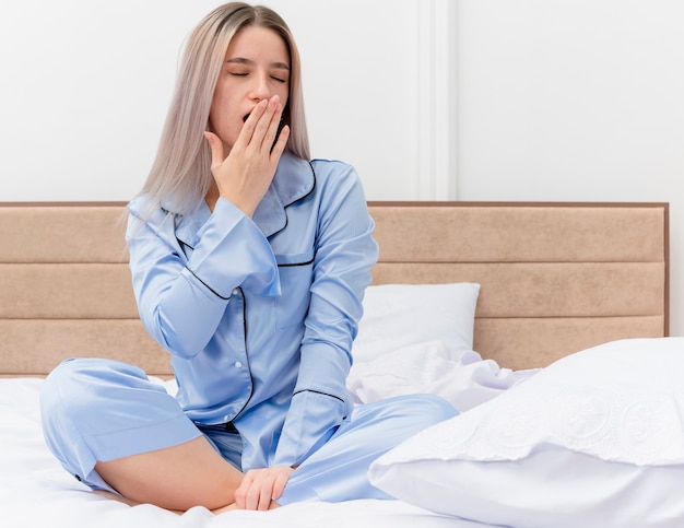 Foto gratuita giovane bella donna in pigiama blu che si siede sul letto nell'interiore della camera da letto su sfondo chiaro