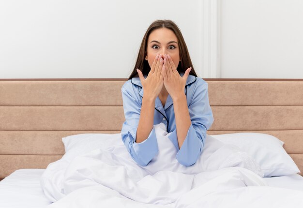 Young beautiful woman in blue pajamas sitting in bed being amazed and surprised in bedroom interior 