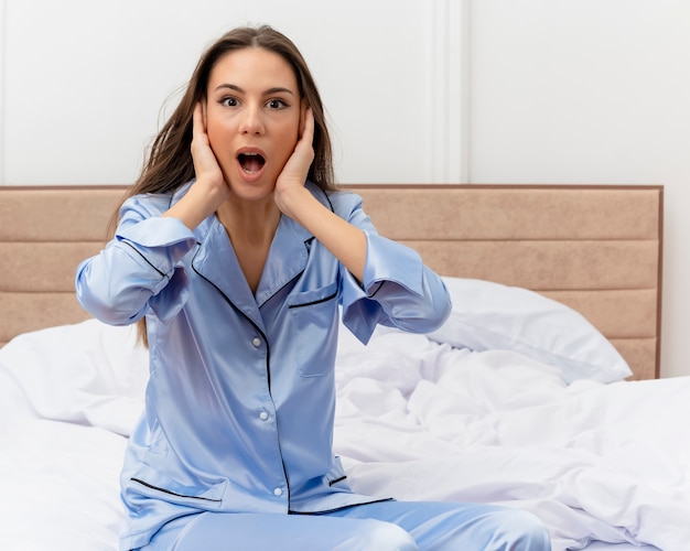 Young beautiful woman in blue pajamas sitting on bed looking at camera being amazed and surprised in bedroom interior on light background