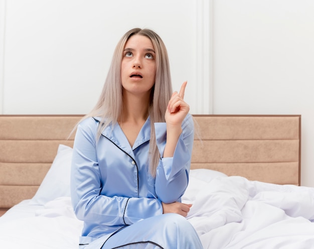 Giovane bella donna in pigiama blu che si siede sul letto che osserva in su che mostra il dito indice nell'interno della camera da letto su sfondo chiaro