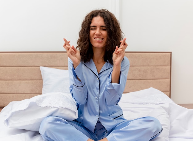 Young beautiful woman in blue pajamas sitting on bed making desirable wish crossing fingers with closed eyes biting lip in bedroom interior on light background