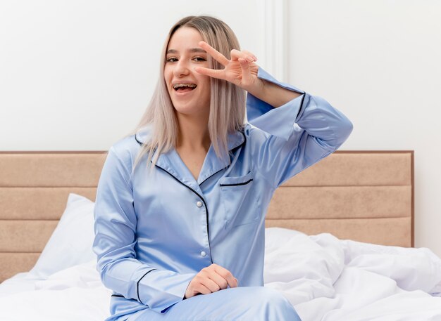 Young beautiful woman in blue pajamas sitting on bed smiling cherrfully showing v-sign in bedroom interior on light background