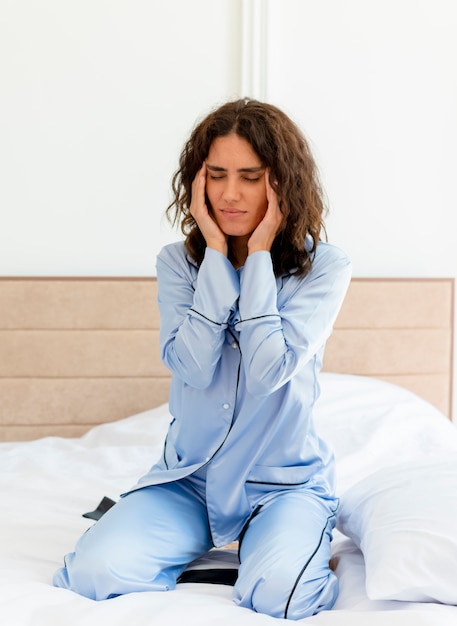 Молодая красивая женщина в синей пижаме сидит на кровати и трогает виски с головной болью в интерьере спальни