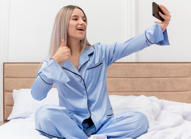 Young beautiful woman in blue pajamas sitting on bed using smartphone doing selfie showing thumbs up in bedroom interior on light background