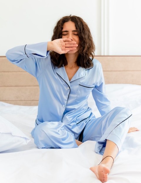 Молодая красивая женщина в синей пижаме сидит на кровати, просыпаясь, чувствуя утреннюю фатугу, зевая в интерьере спальни на светлом фоне
