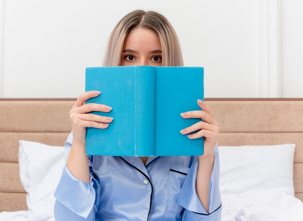 Young beautiful woman in blue pajamas sitting on bed with book hiding face peeking over in bedroom interior on light background