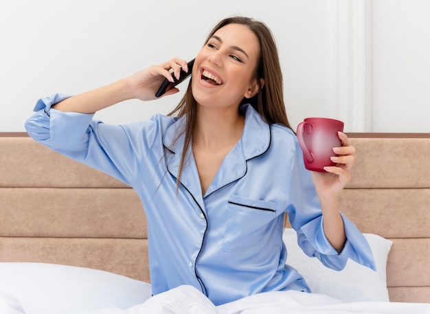 Giovane bella donna in pigiama blu che si siede sul letto con la tazza di caffè che tiene smartphone che guarda l'obbiettivo felice ed emozionato parlando al telefono cellulare all'interno della camera da letto su sfondo chiaro