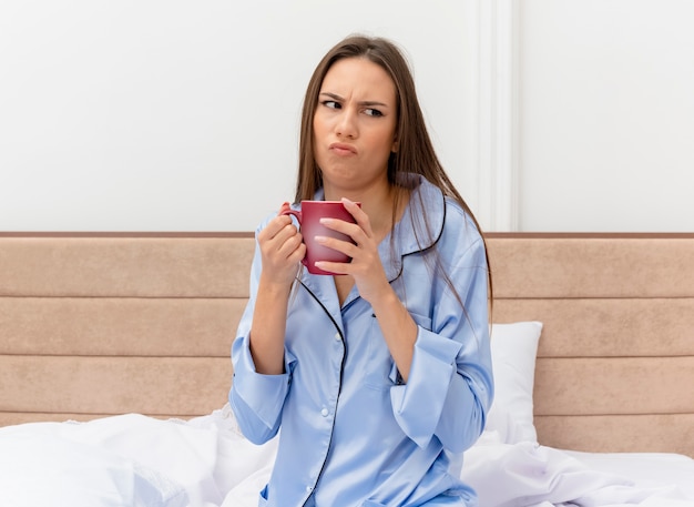 Young beautiful woman in blue pajamas sitting on bed with cup of coffee looking aside being displeased in bedroom interior on light background