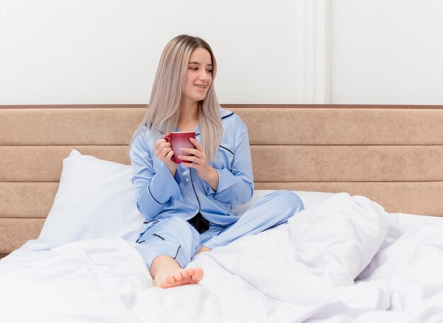 Молодая красивая женщина в синей пижаме сидит на кровати с чашкой кофе, глядя в сторону, улыбаясь, отдыхая, наслаждаясь утренним временем в интерьере спальни