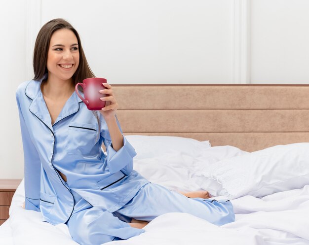 Young beautiful woman in blue pajamas sitting on bed with cup of coffee looking aside with smile on face resting in bedroom interior on light background