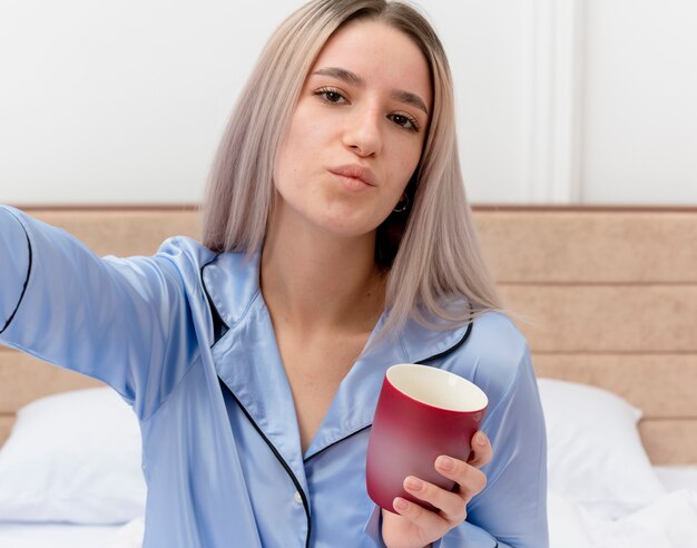 Young beautiful woman in blue pajamas sitting on bed with cup of coffee looking at camera smiling confident in bedroom interior on light background