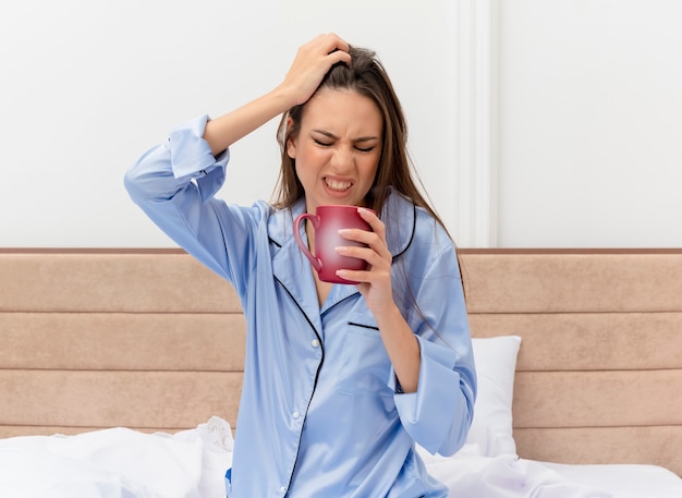 Young beautiful woman in blue pajamas sitting on bed with cup of coffee looking with annoyed expression waking up in bedroom interior on light background