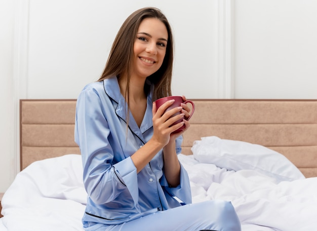 Young beautiful woman in blue pajamas sitting on bed with cup of coffee resting looking at camera happy and positive smiling enjoying weekend in bedroom interior on light background